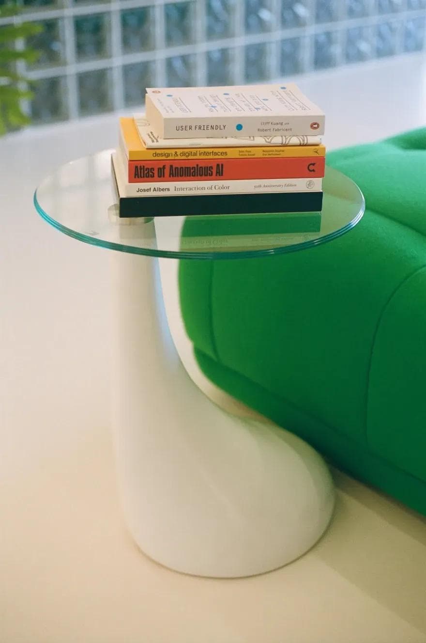 A round, glass table with a stack of colourful design books on top, next to a green couch. 