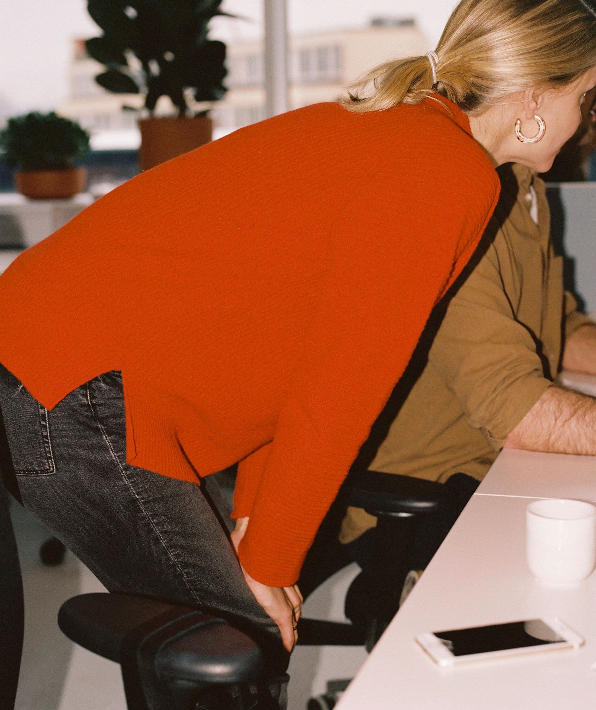 Woman wearing a red sweater, looking with her hands on her knees at a colleague's screen. 