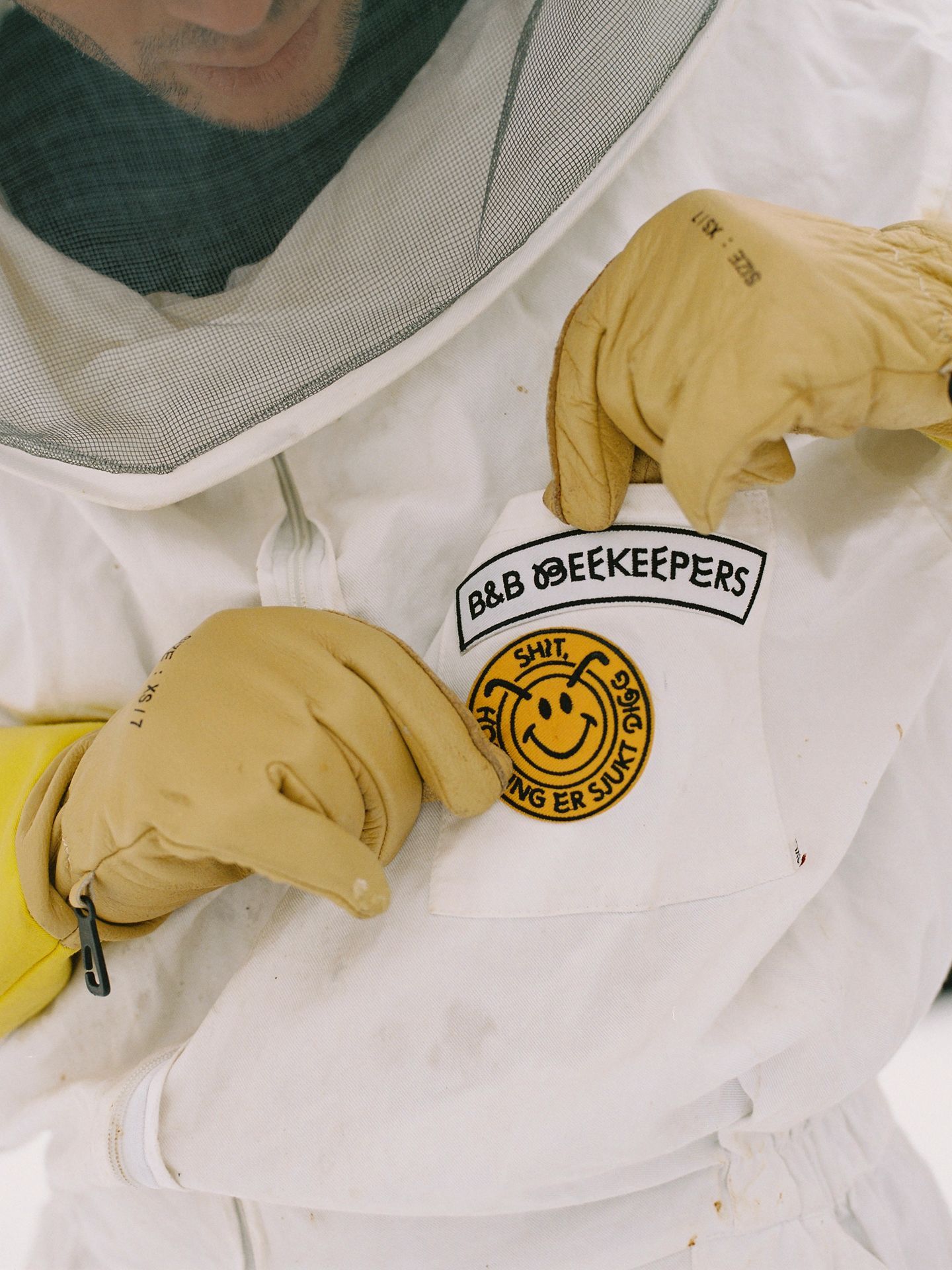 Photo of a beekeeper wearing yellow gloves, who is looking at the BB-branded patches on his suit that say "B&B Beekeepers" and "Save the Bees!".