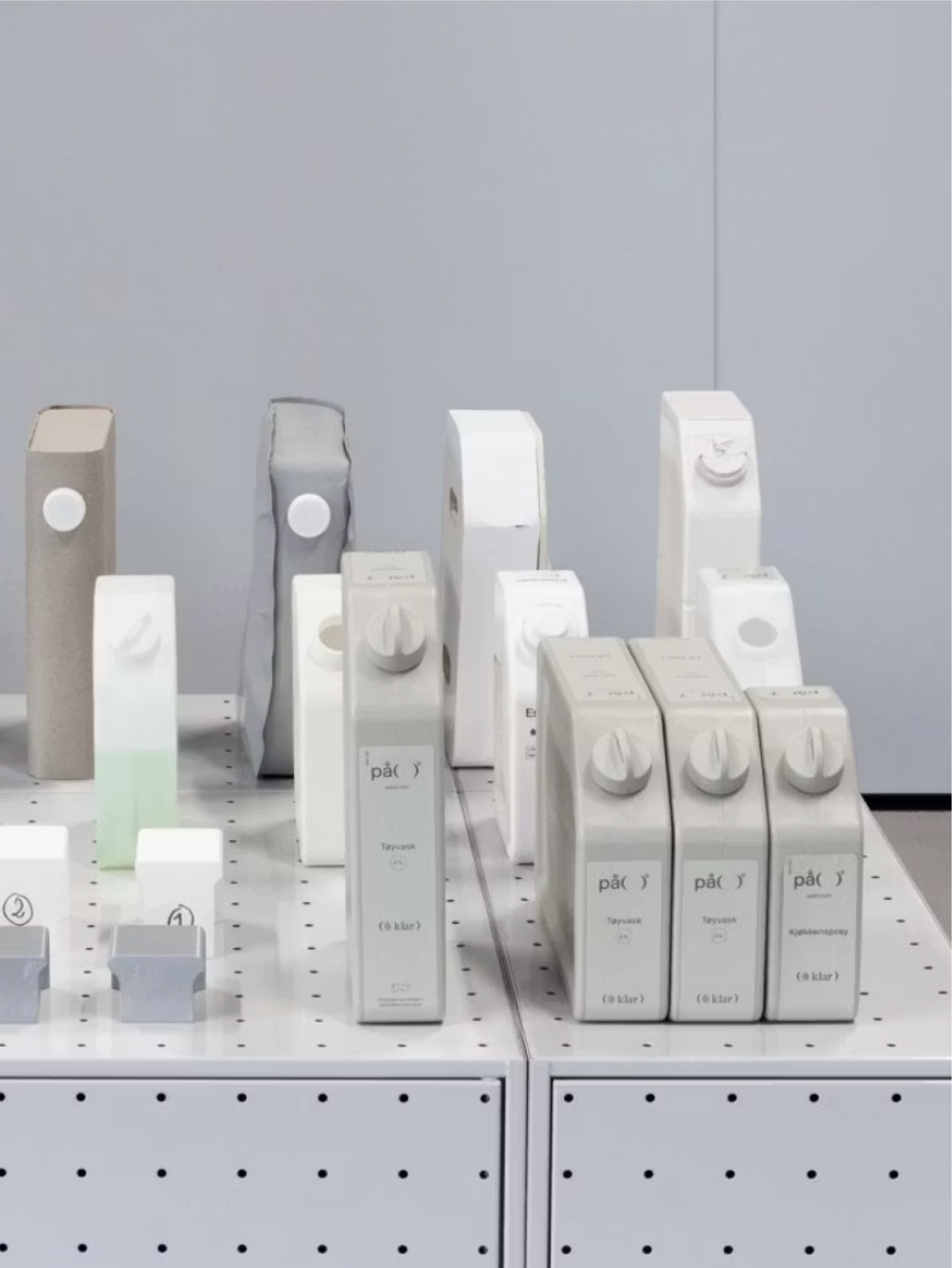 A display of minimalist, refillable household product canisters in neutral tones on a white grid shelf.