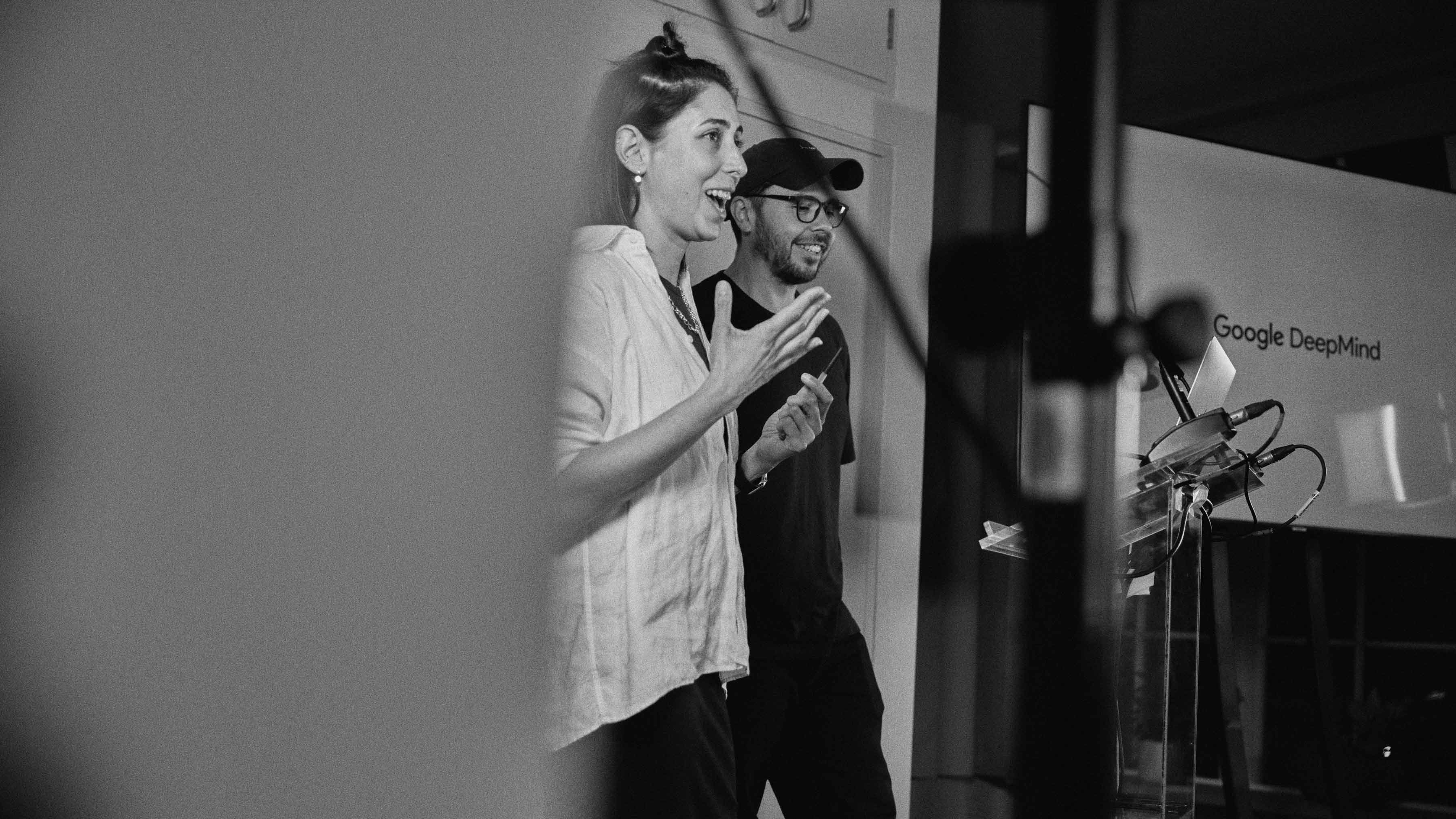 Black and white photo of two of the speakers on stage, smiling, with the Google Deepmind logo on screen.