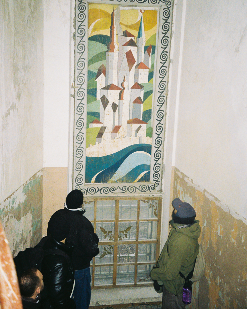 Two men stand in a staircase, looking at an old painting in a wall.