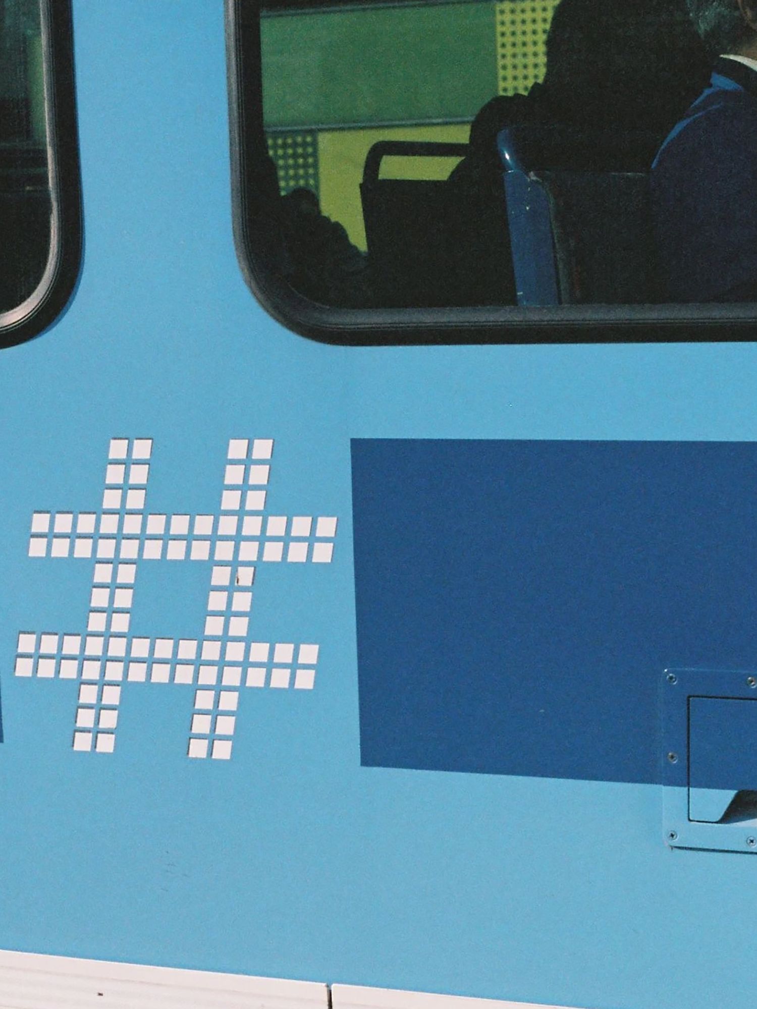 Close-up of the side of a tram showing the Ruter logo. The logo is a hashtag consisting out of white squares. The tram is light blue with a darker blue horizontal line beneath the windows