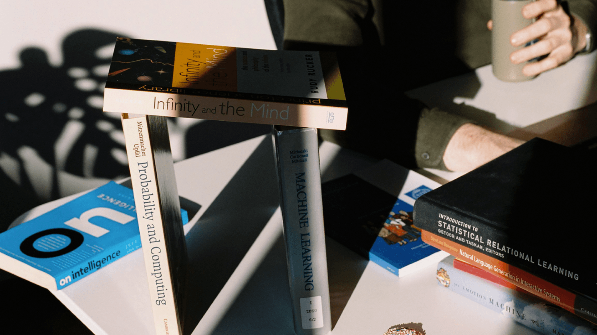 Image of various books on computer science and math, stacked in a playful manner on a desk. Behind it, someone is working while holding a tumbler.