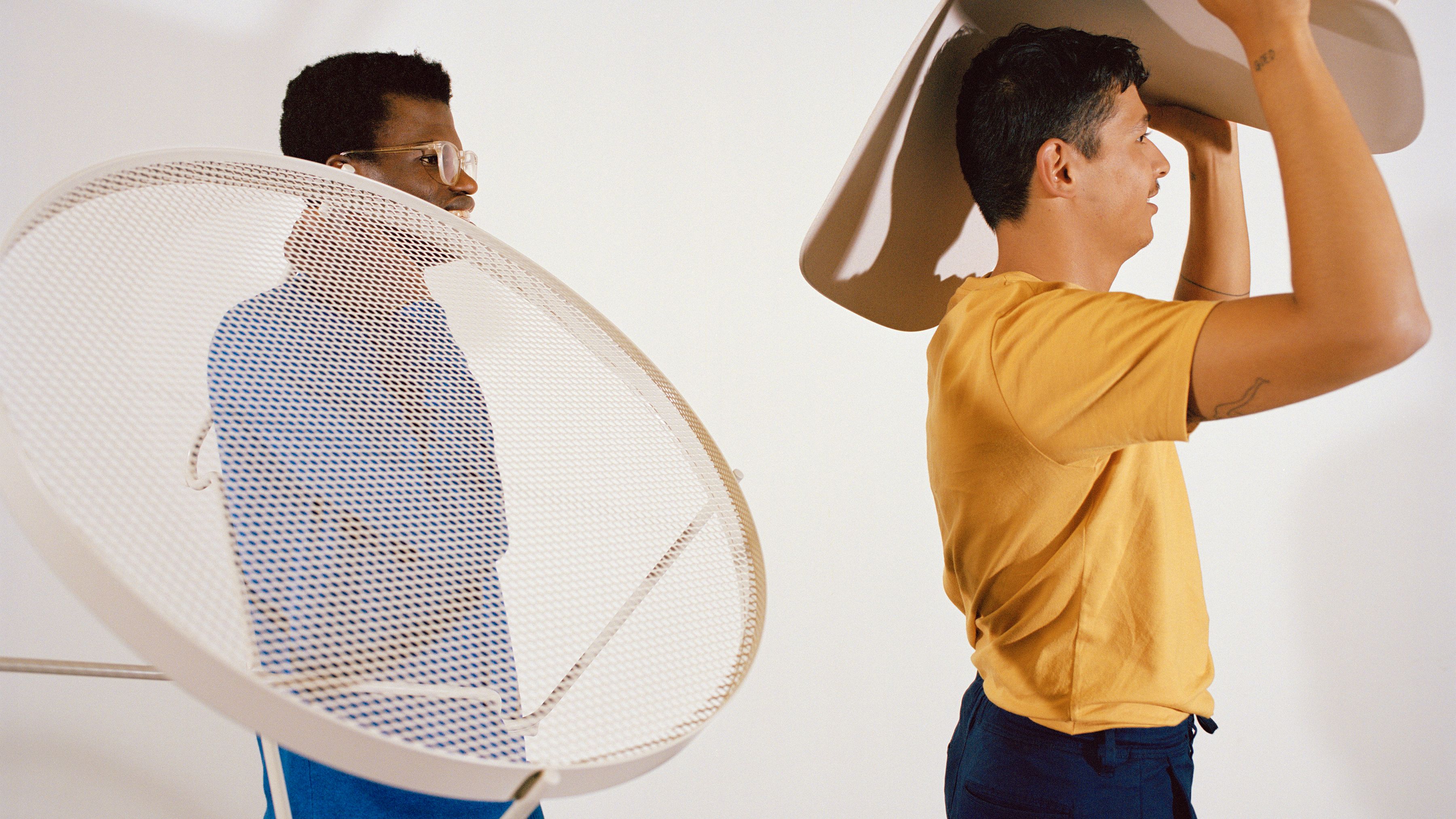 Two men carrying furniture. One wears a blue sweater and carries a white table on his shoulder. The other wears a yellow t-shirt and a beige chair above his head.