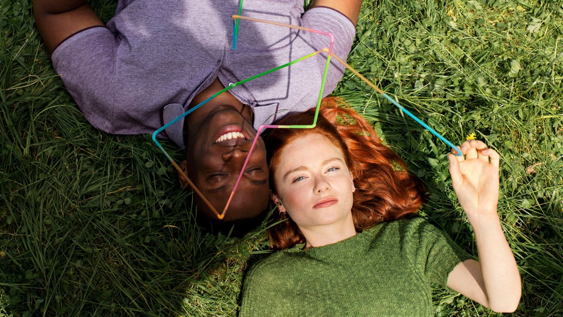 Two smiling people laying on the grass, holding plastic straws that form a colourful graph.