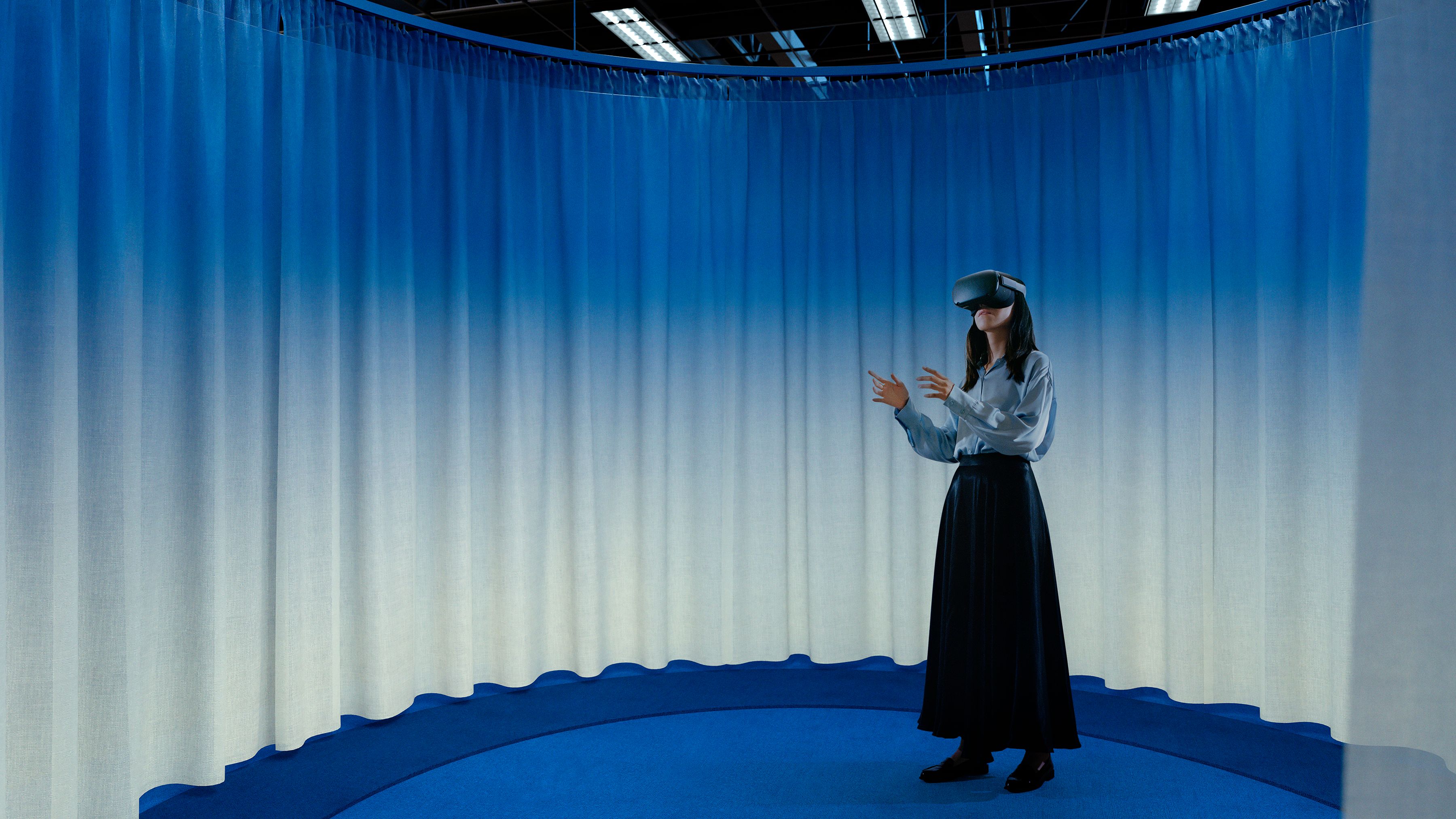 Woman standing in a round space surrounded by blue curtains. She is wearing VR-goggles and has her hands in mid-air.