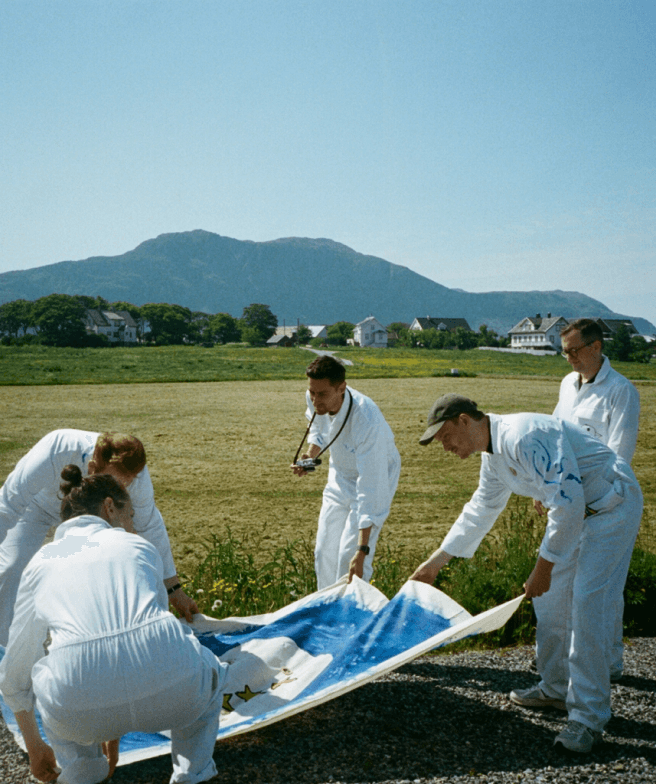 People in white coats holding laying down a large painted flag. The people are outside in a large field with a mountain in the background