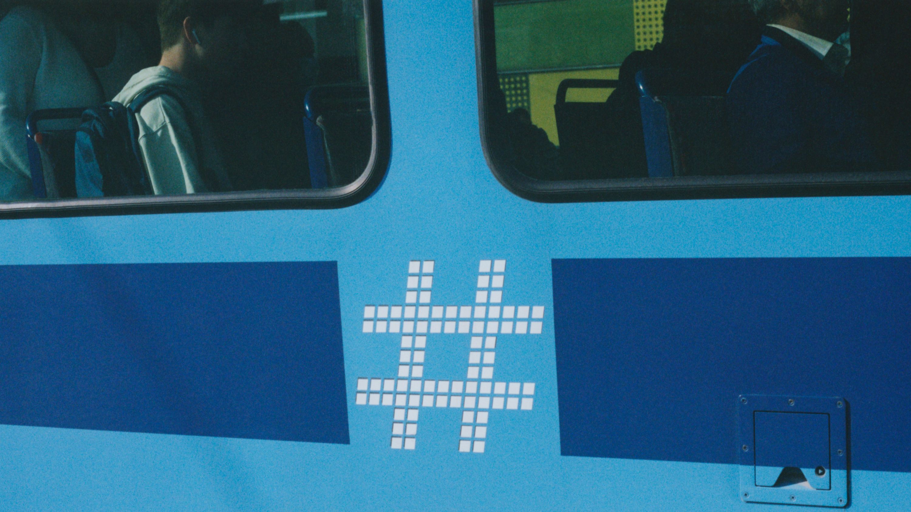 Close-up of the side of a tram showing the Ruter logo. The logo is a hashtag consisting out of white squares. The tram is light blue with a darker blue horizontal line beneath the windows