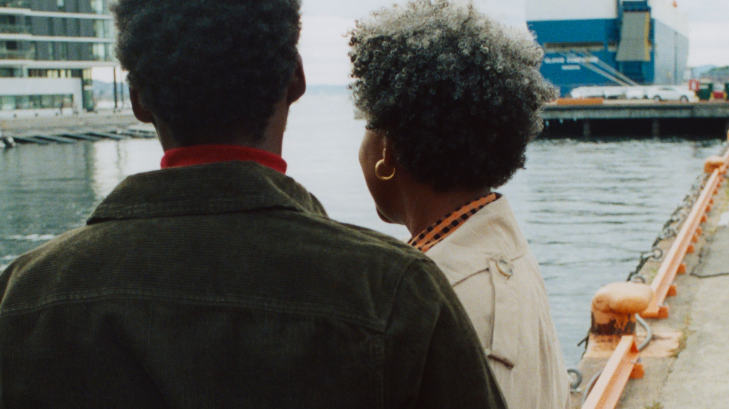 Close-up photo from the back of two people at a harbor in an industrial area, looking out over the water