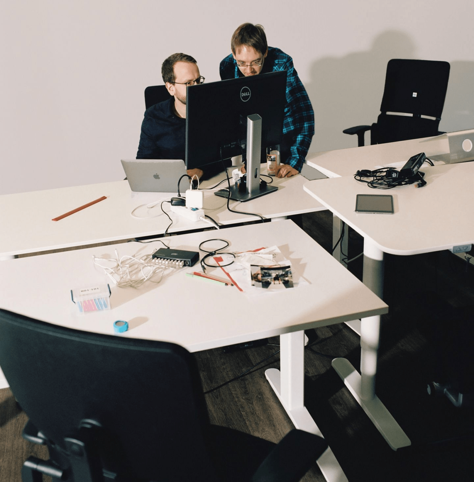 Two mean sitting and standing behind desk, looking at a screen. On the desk, there are many cables and chargers, as well as pencils, books and other office supplies.